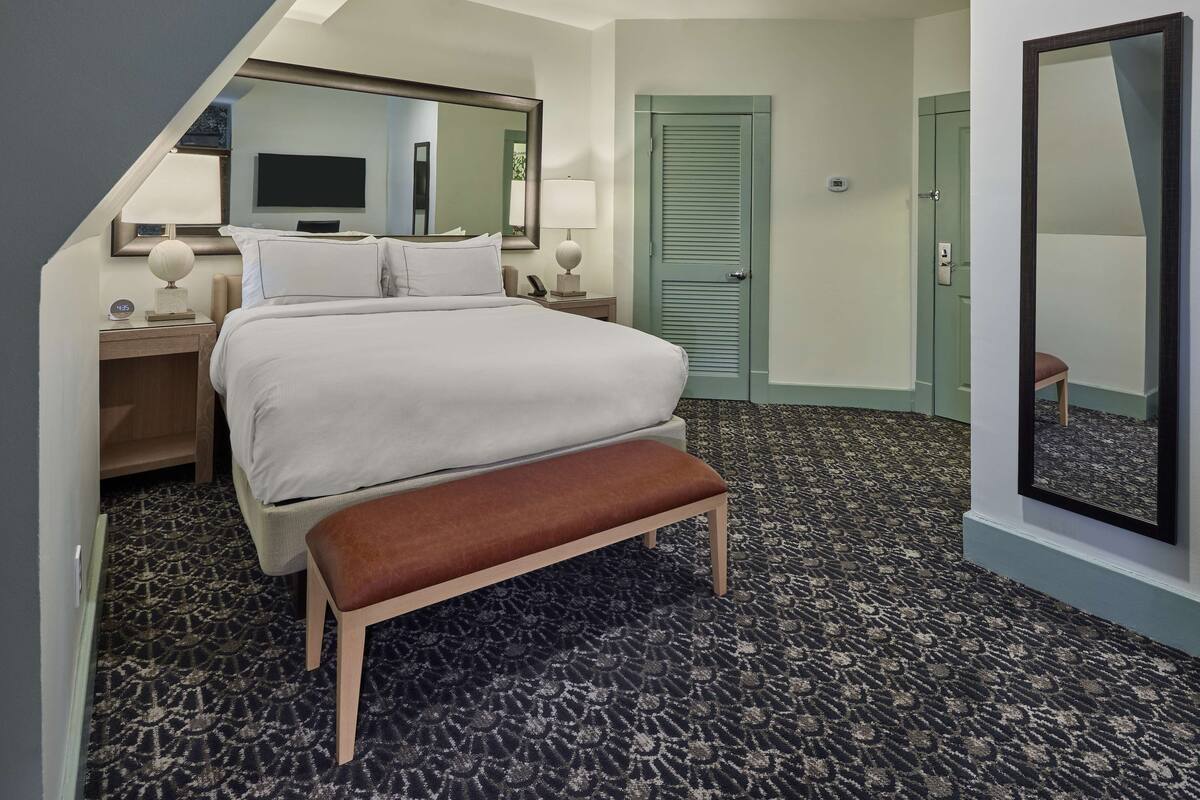 A hotel room with a large bed, a bench, a desk with a lamp, a mirror, and a door.