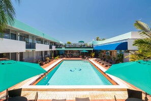 A pool area with lounge chairs and umbrellas, surrounded by a building with balconies and a covered walkway.