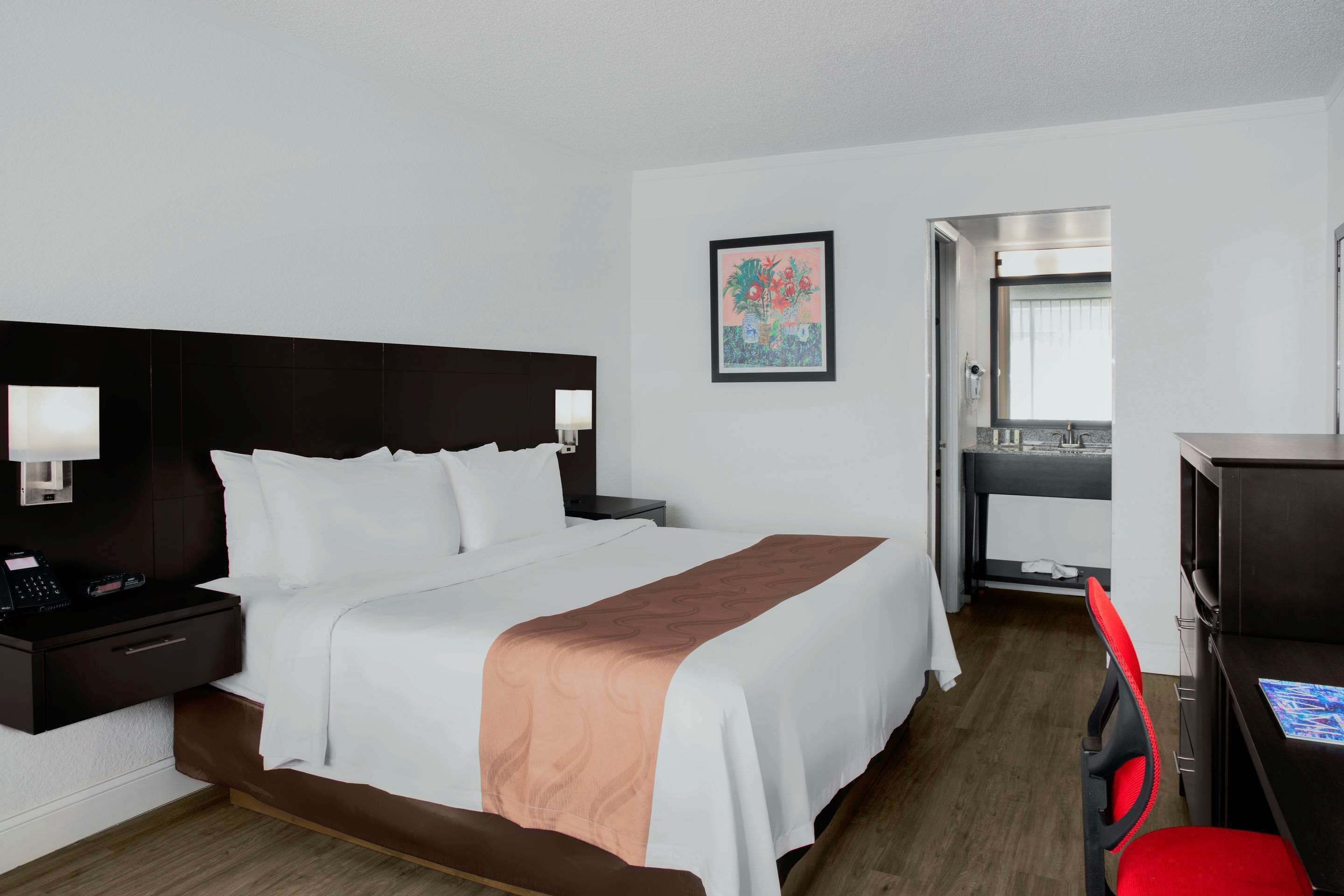 Ein Hotelzimmer mit Bett, Nachttisch, Lampe, Schreibtisch, Stuhl und einem gerahmten Bild an der Wand.