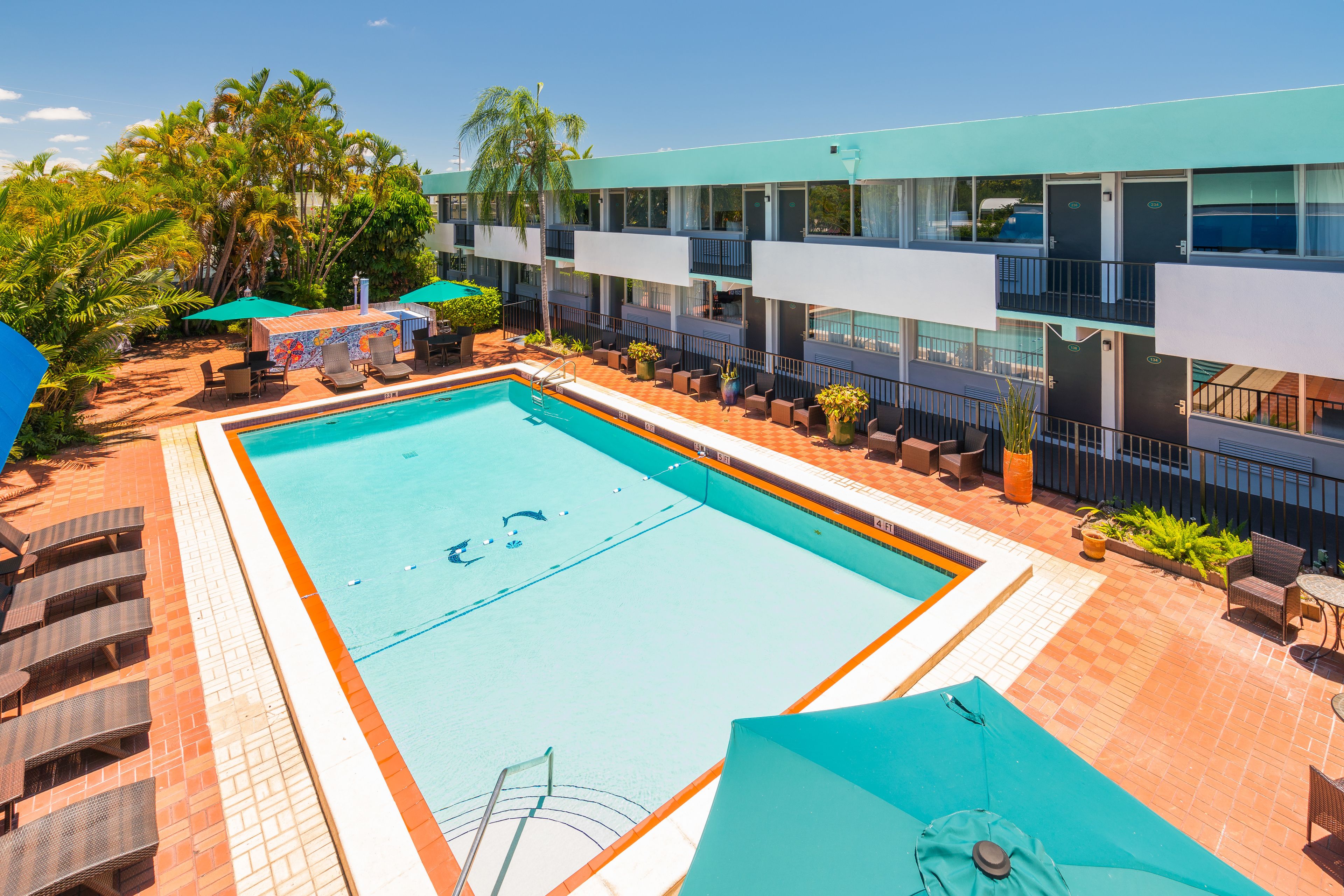 Una piscina con sillones de descanso y sombrillas, rodeada por un edificio con balcones y un patio empedrado.