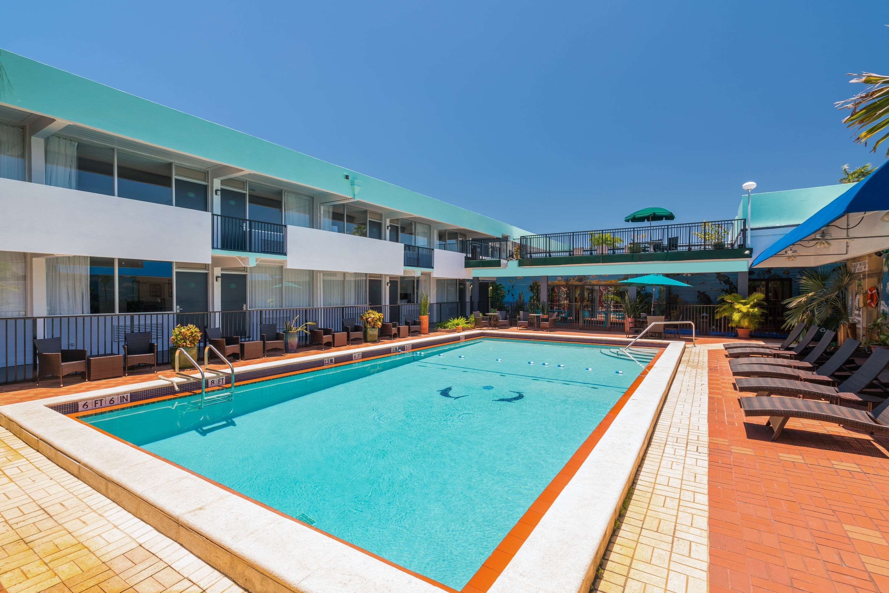 Una piscina con sillas de descanso y una zona de estar cubierta, rodeada por un edificio de varios pisos con balcones.