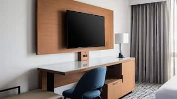 A hotel room with a flat-screen TV mounted on a wooden panel, a desk with a chair, a bedside lamp, and a large window with curtains.
