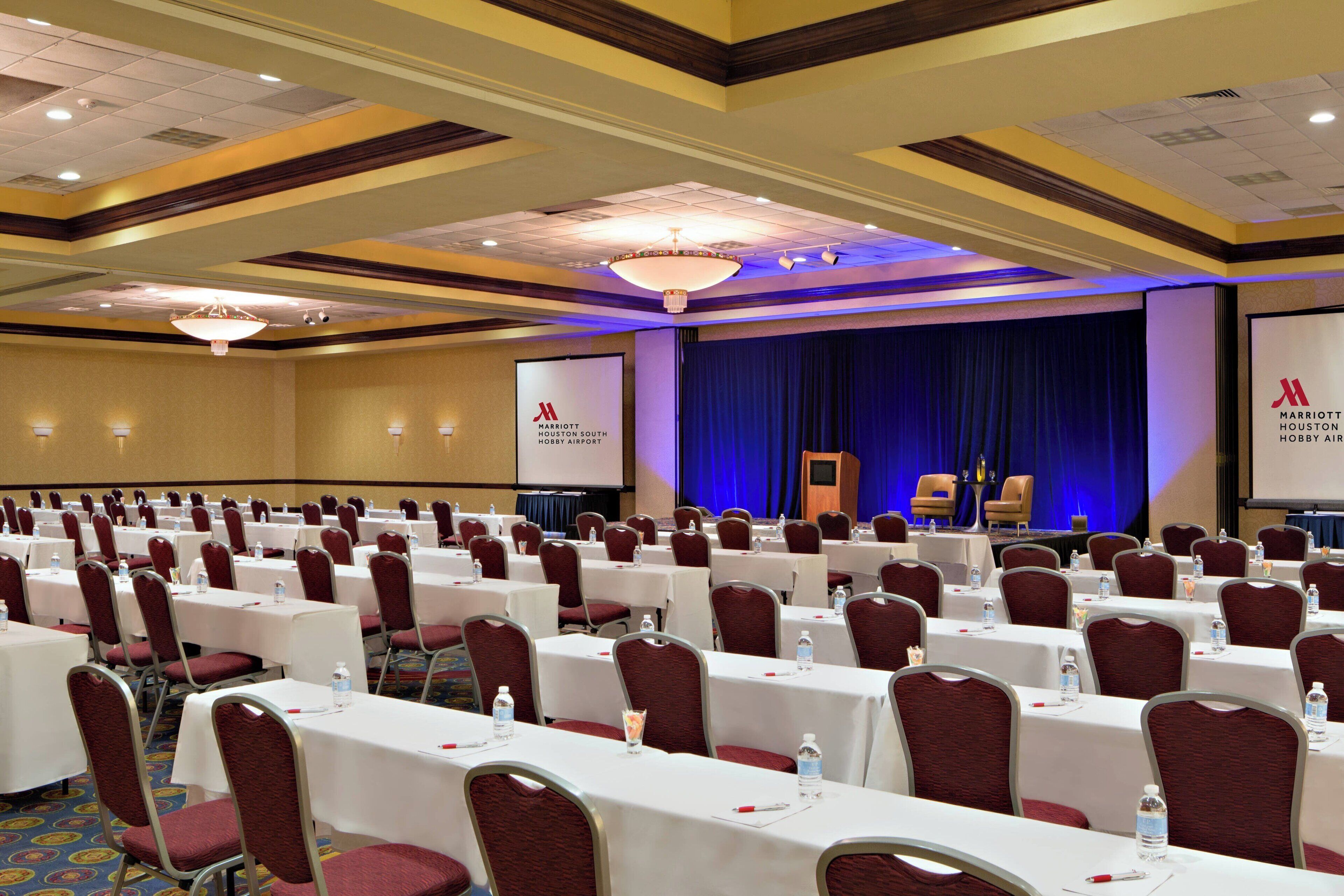 Een congreszaal met rijen tafels en stoelen, een podium met een blauw gordijn, en een scherm waarop het Marriott-logo wordt getoond.