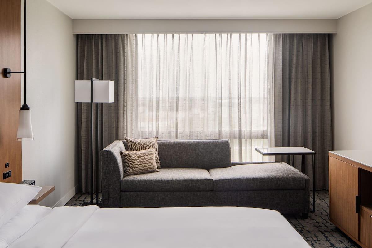 A hotel room with a bed, a sofa, a small table, a lamp, and a window with curtains.