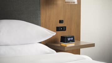 Habitación de hotel con una mesita de noche de madera, un reloj digital que marca las 12:48 y una lámpara en la pared.