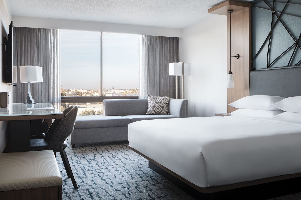 A modern hotel room with a large bed, a desk with a chair, a sofa, and a view of the city through large windows.