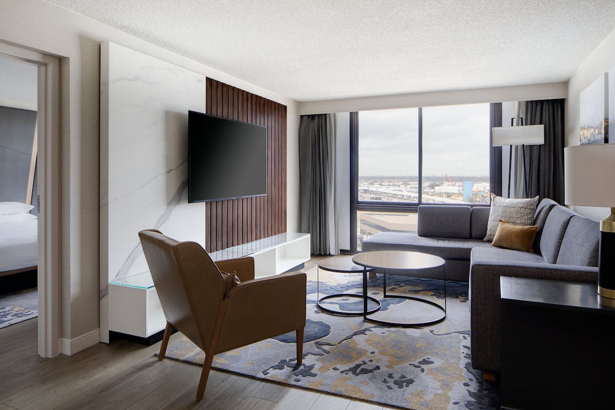 A modern hotel room with a large TV, a sofa, a chair, and a view of the city.
