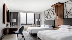 Habitación de hotel con dos camas, un escritorio, una silla, un televisor y vistas a la ciudad.