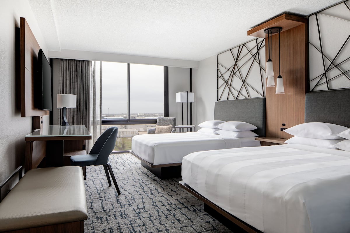A hotel room with two beds, a desk, a chair, a TV, and a view of the city.