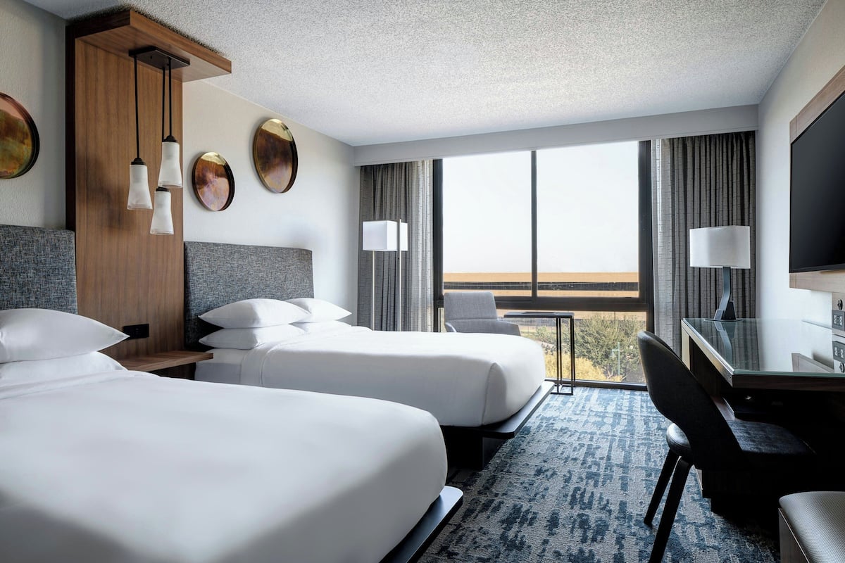 A hotel room with two beds, a desk, a chair, a TV, and a large window with a view.