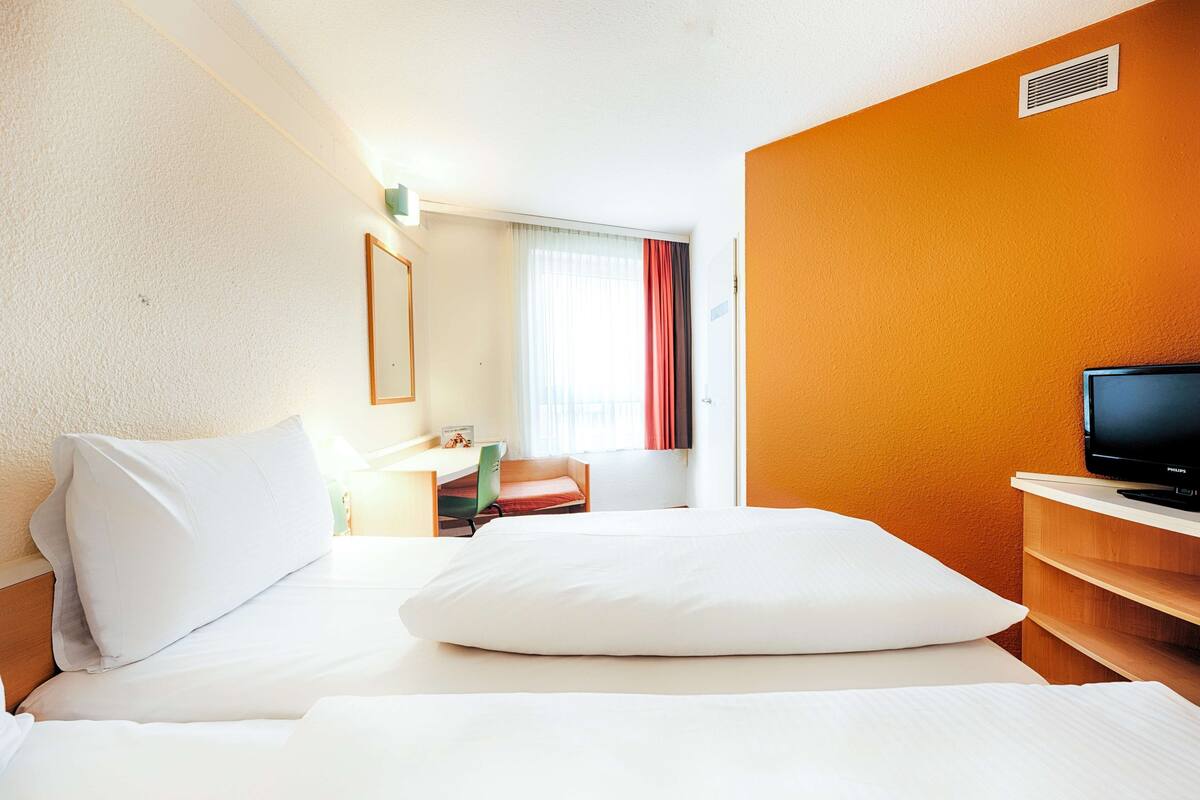 Une chambre d’hôtel avec un lit, un bureau, une chaise, une télévision et une fenêtre avec des rideaux.