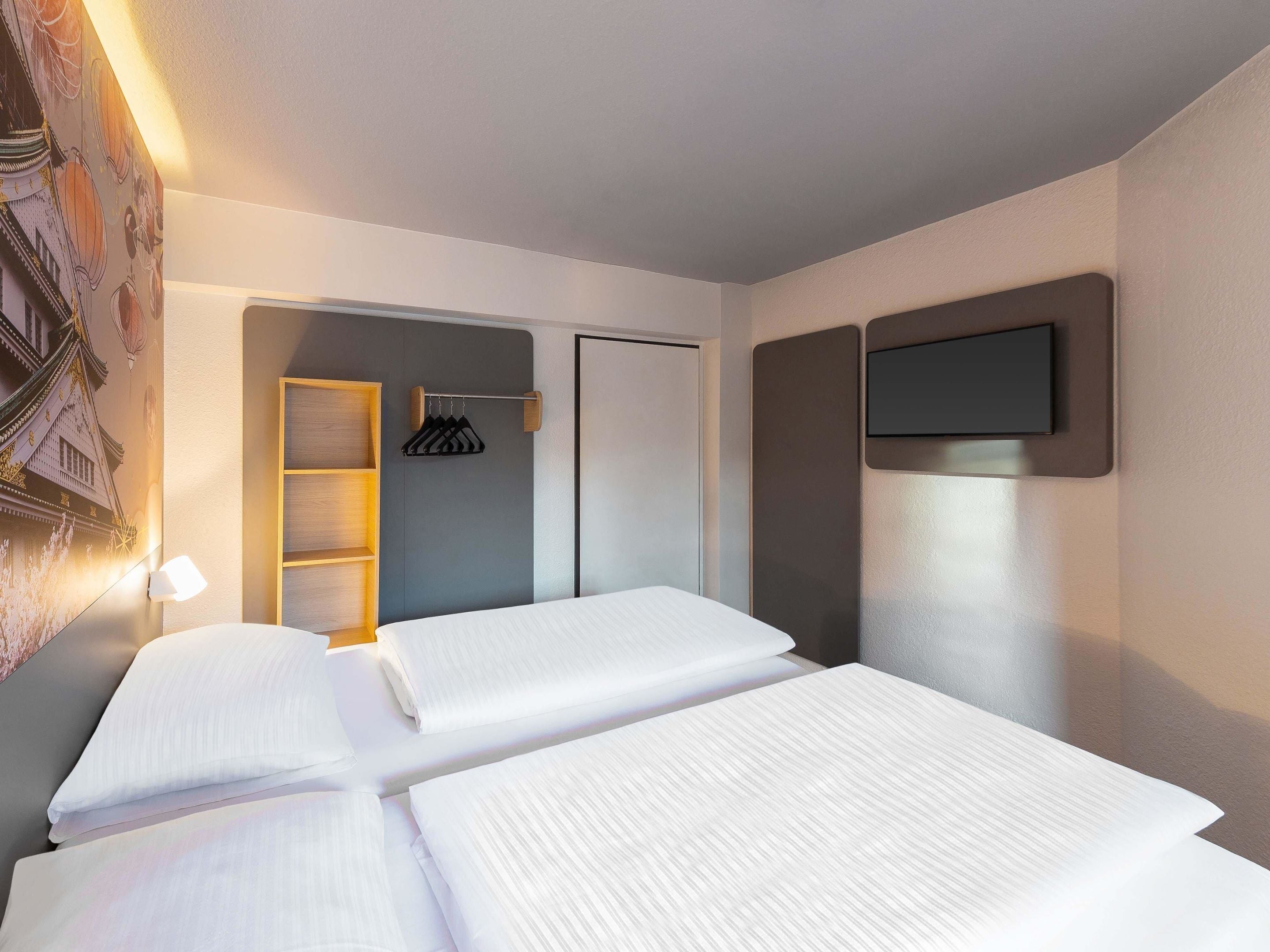 Ein Hotelzimmer mit zwei Betten, einem Kleiderschrank, einem an der Wand befestigten Fernseher und einem Wandregal mit Lampe.