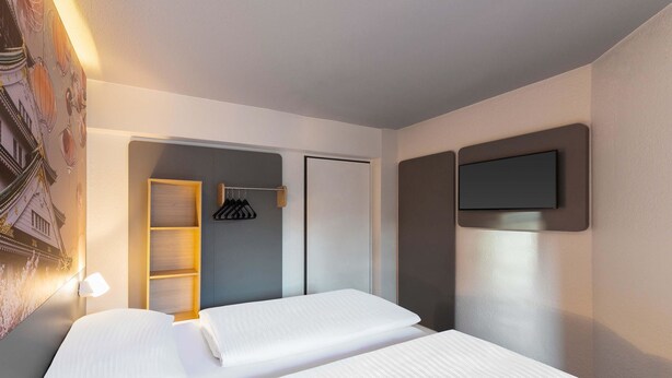 A hotel room with two beds, a wardrobe, a TV mounted on the wall, and a wall-mounted shelf with a lamp. - B&B Hotel Düsseldorf-Hbf (Düsseldorf)