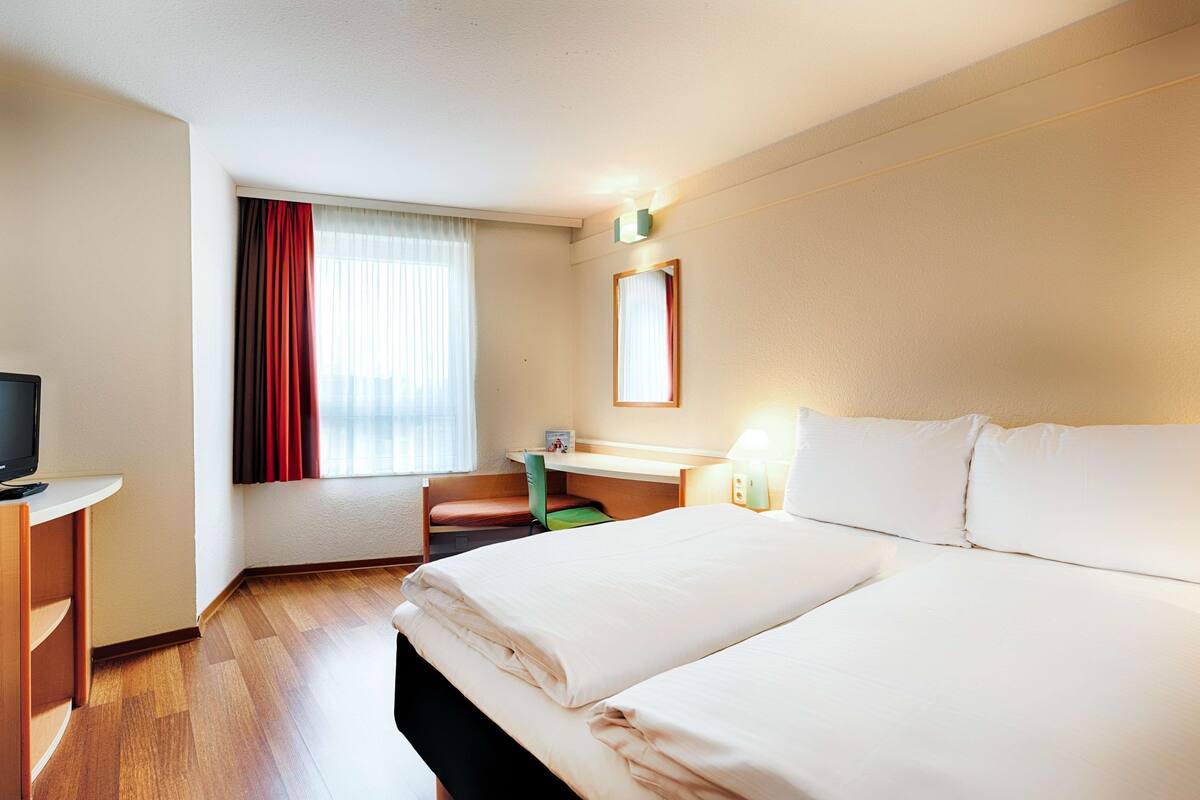 Une chambre d’hôtel avec deux lits, un bureau, une chaise, une télévision et une fenêtre avec des rideaux.