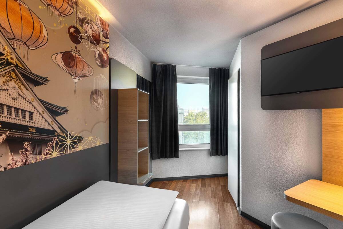 Une chambre d’hôtel moderne, équipée d’un lit, d’une télévision, d’une fenêtre avec des rideaux et d’un mur au décor traditionnel.