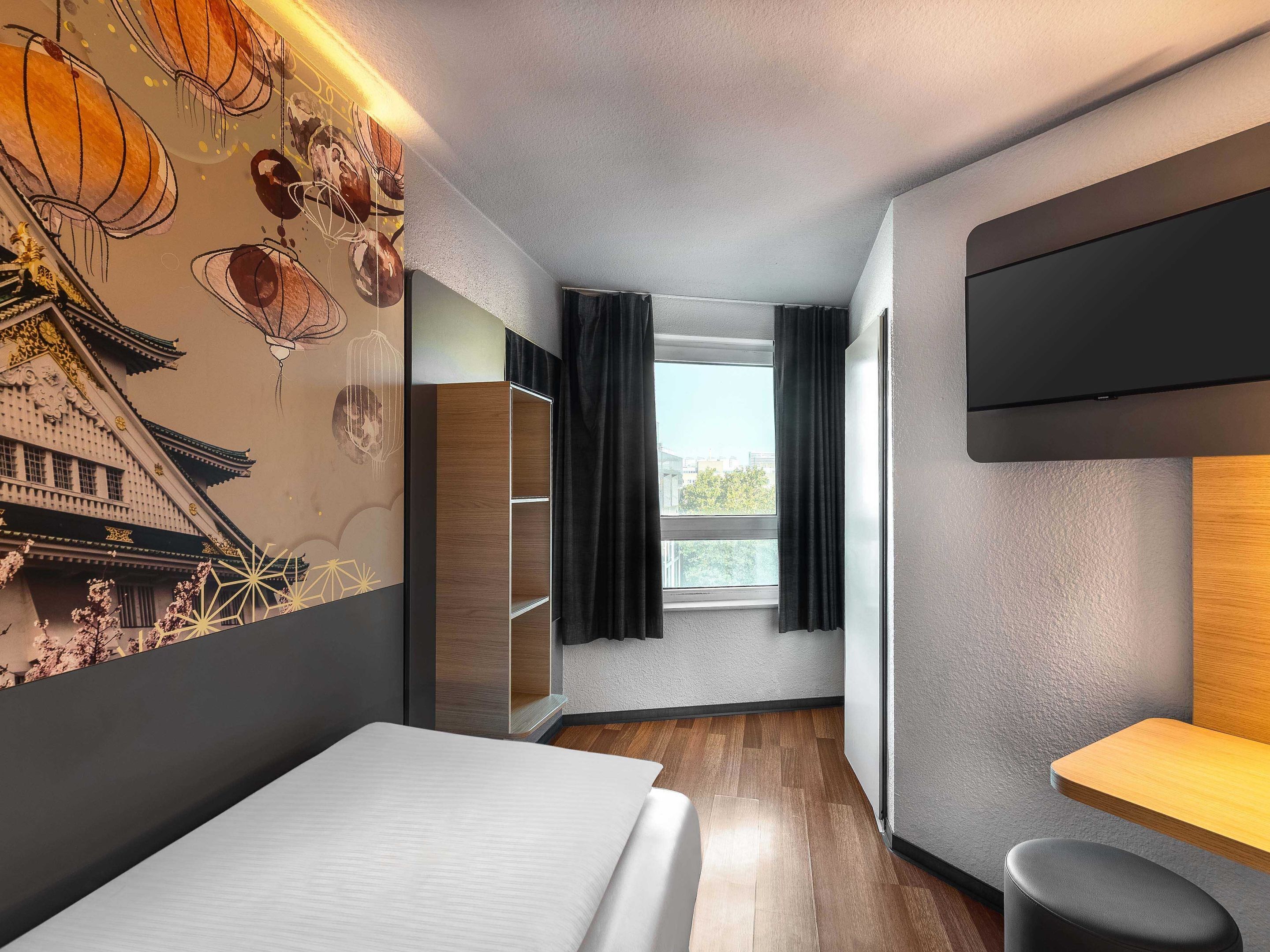 Une chambre d’hôtel moderne, équipée d’un lit, d’une télévision, d’une fenêtre avec des rideaux et d’un mur au décor traditionnel.