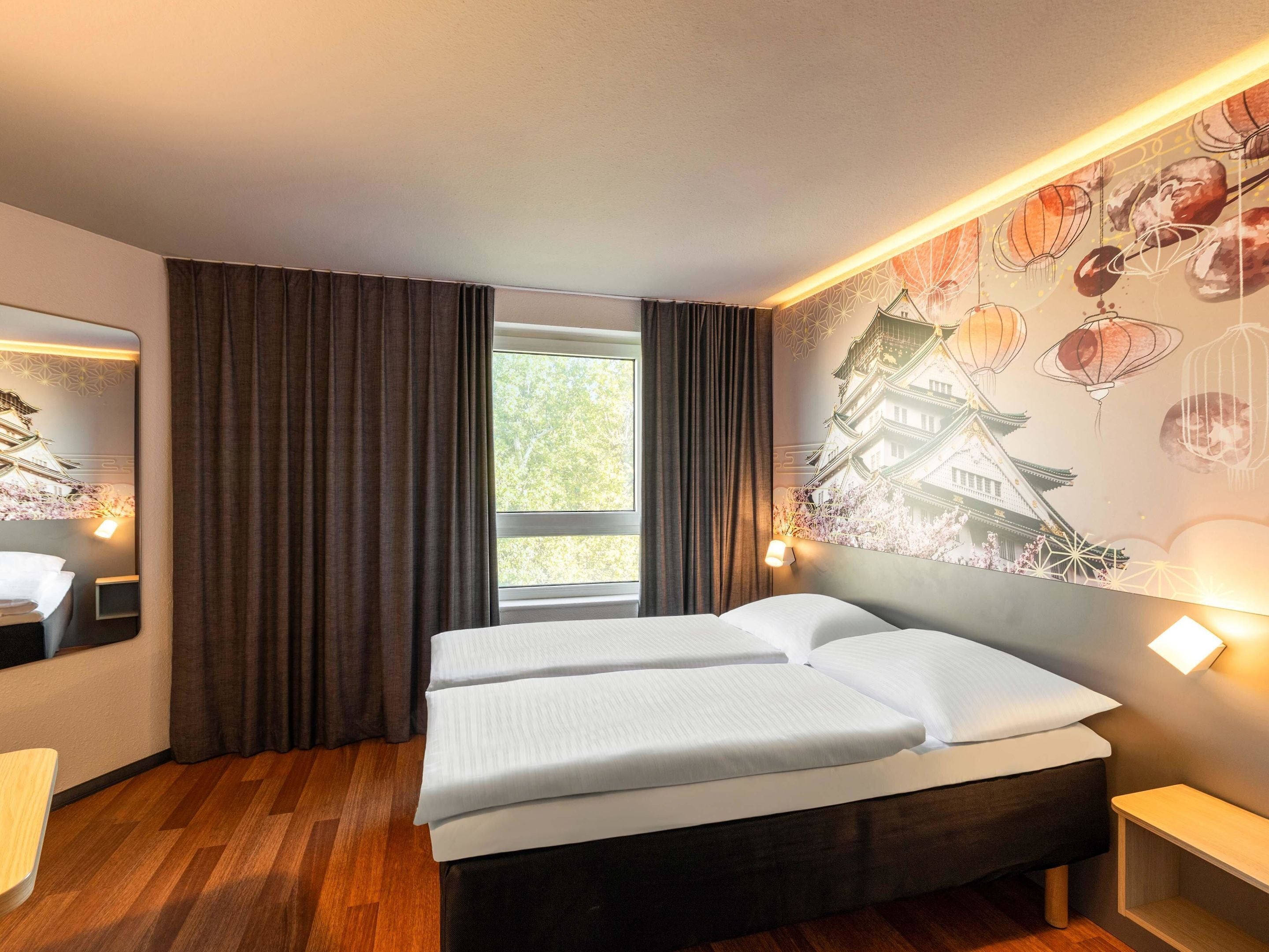 Habitación de hotel con una cama, ventana con cortinas, lámpara de pared y un mural decorativo.