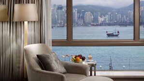 A hotel room with a view of a city skyline across a body of water, a comfortable armchair, a small table with fruit, and a lamp.