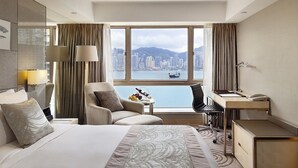 A hotel room with a bed, a desk, a chair, and a view of a cityscape through large windows.
