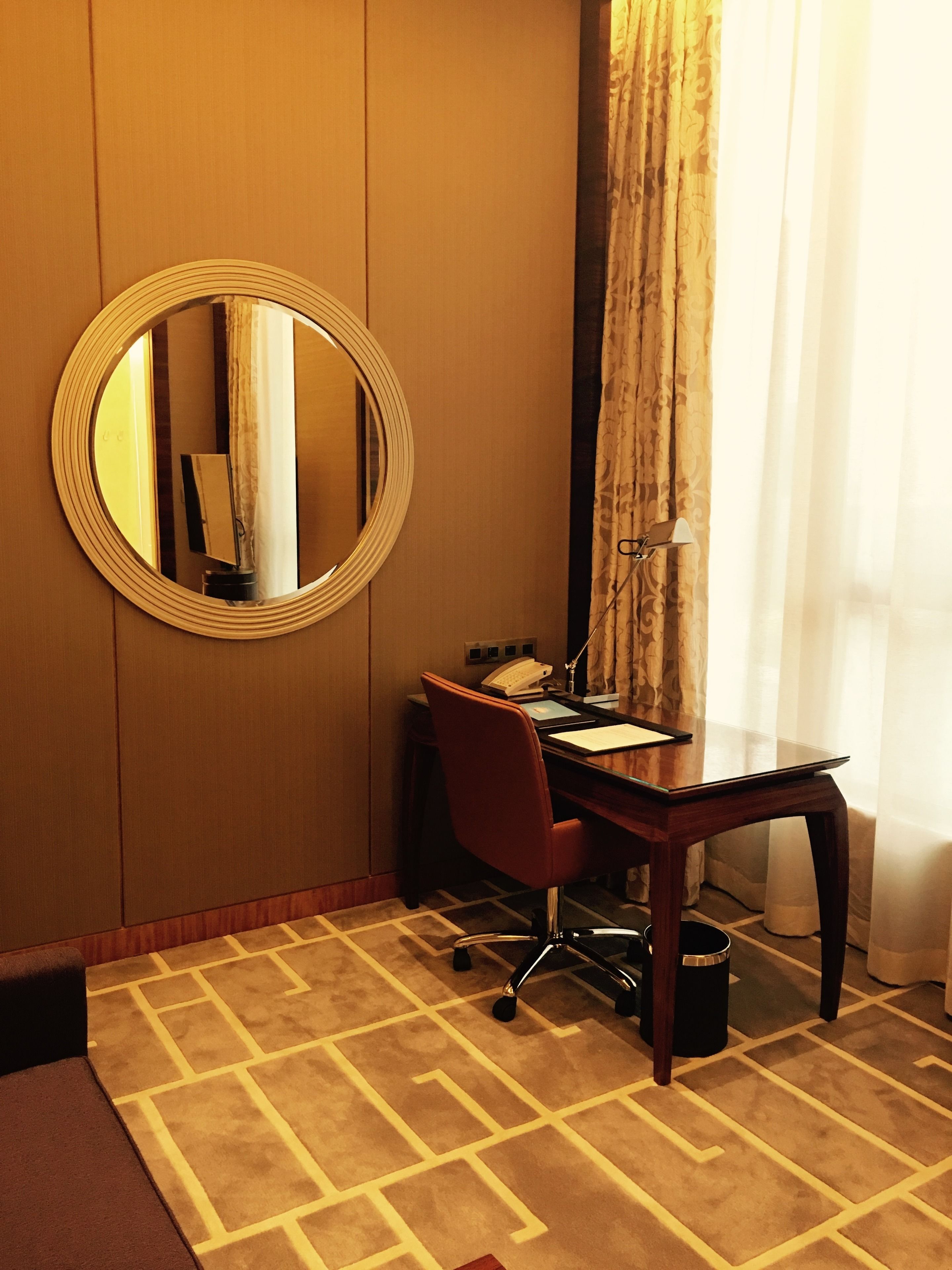 A hotel room with a wooden desk, a red chair, a round mirror, a telephone, and a lamp.