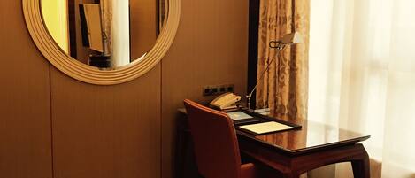 A hotel room with a wooden desk, a red chair, a round mirror, a telephone, and a lamp.