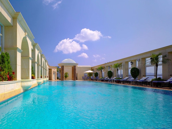 A large swimming pool with lounge chairs and umbrellas, surrounded by a building with columns and a dome structure.