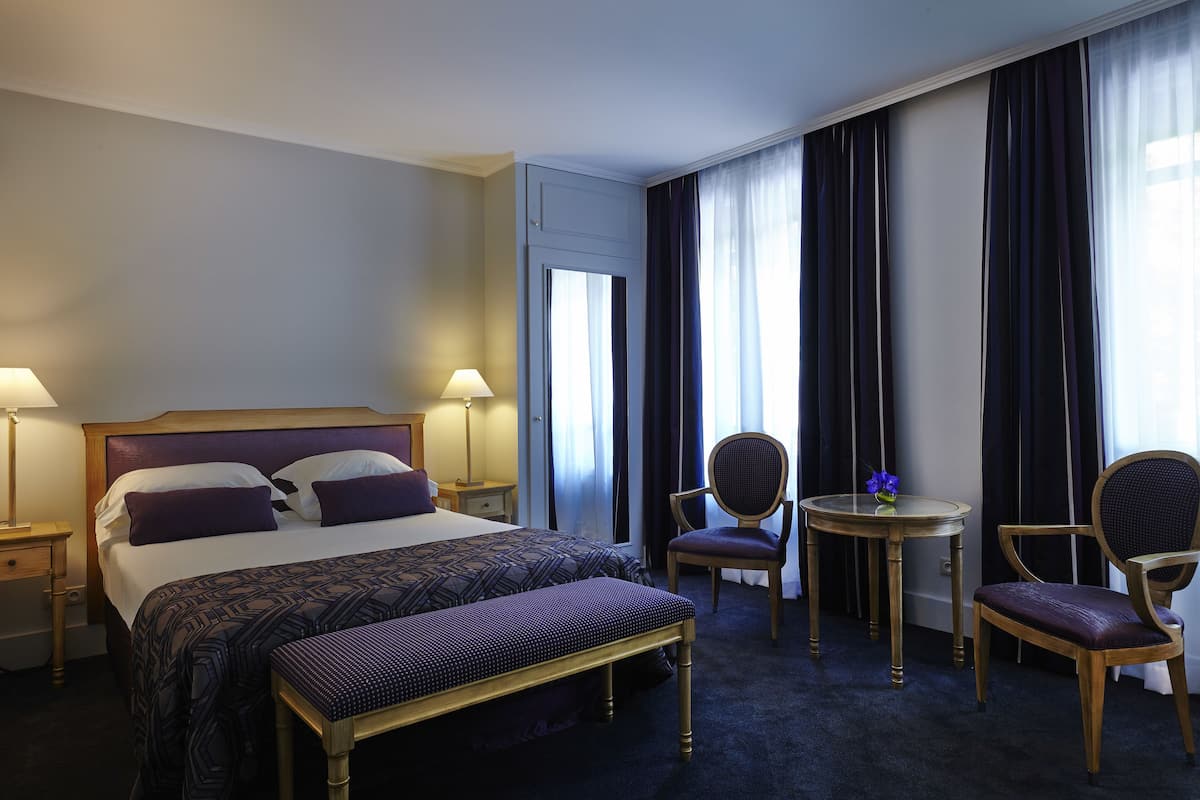 Une chambre d’hôtel avec un grand lit, deux chaises, une petite table et un banc.