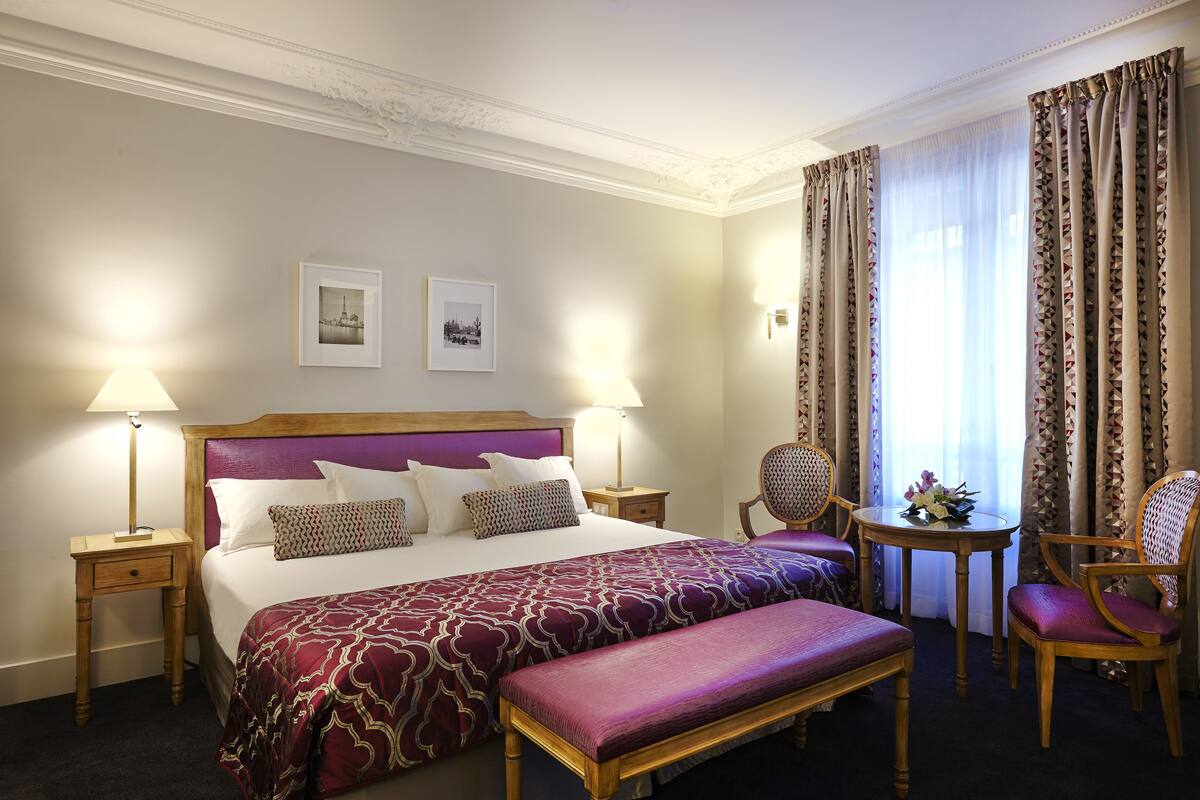 Une chambre d’hôtel avec un grand lit, deux tables de chevet, un banc, une chaise et une petite table agrémentée d’un arrangement floral.