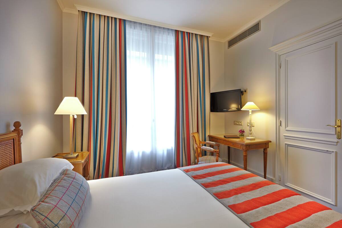 Une chambre d’hôtel comprenant un lit, deux lampes de chevet, un bureau avec une chaise, une télévision et une porte.