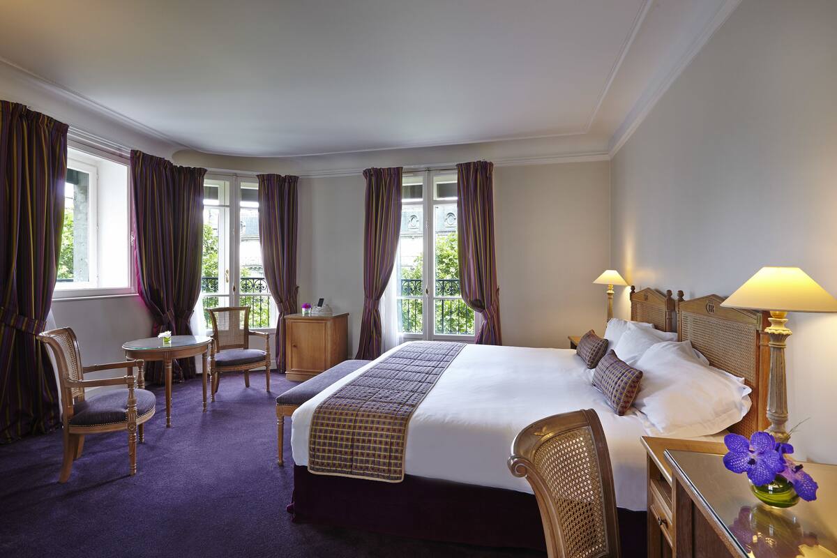 Une chambre d’hôtel avec un grand lit, deux chaises, une petite table et une table de chevet avec une lampe.