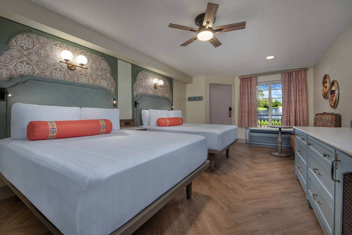 A hotel room with two beds, a ceiling fan, a window with curtains, and a small table.