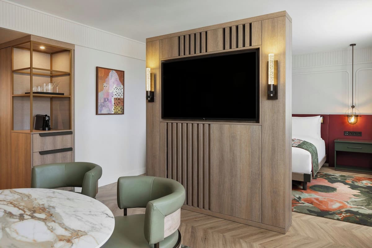 Habitación de hotel con pared de paneles de madera, televisor de pantalla plana, cama, sofá verde, mesita de mármol y estantería con objetos decorativos.
