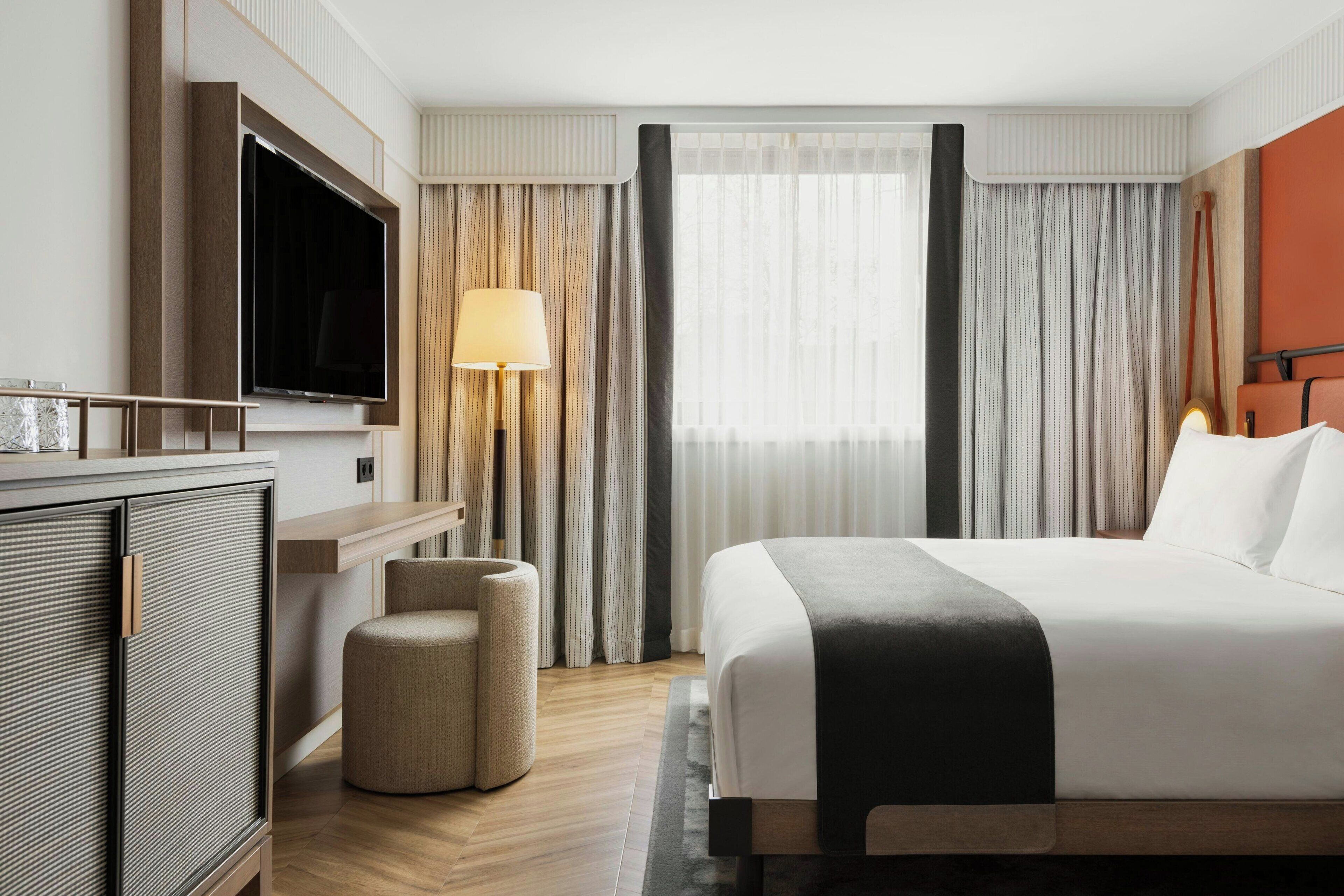 Quarto de hotel com uma cama grande, televisão fixada na parede, área de estar com uma poltrona e apoio para os pés, e janela com cortinas.
