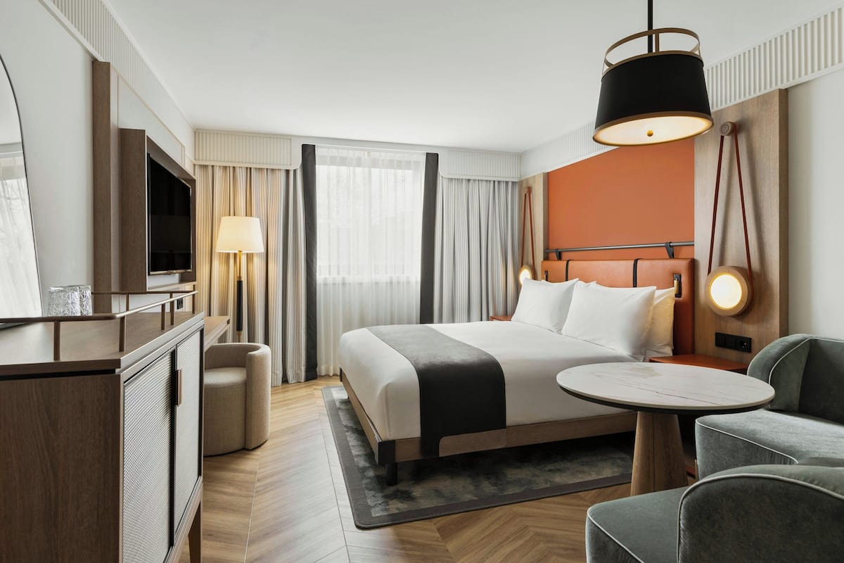 Una habitación de hotel moderna con cama, mesita de noche, lámpara y una zona de estar con una silla y una pequeña mesa.