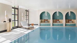 Une piscine intérieure moderne avec une eau d’un bleu cristallin, un espace détente avec des chaises blanches et un coin salon doté de bancs rembourrés et de coussins décoratifs.