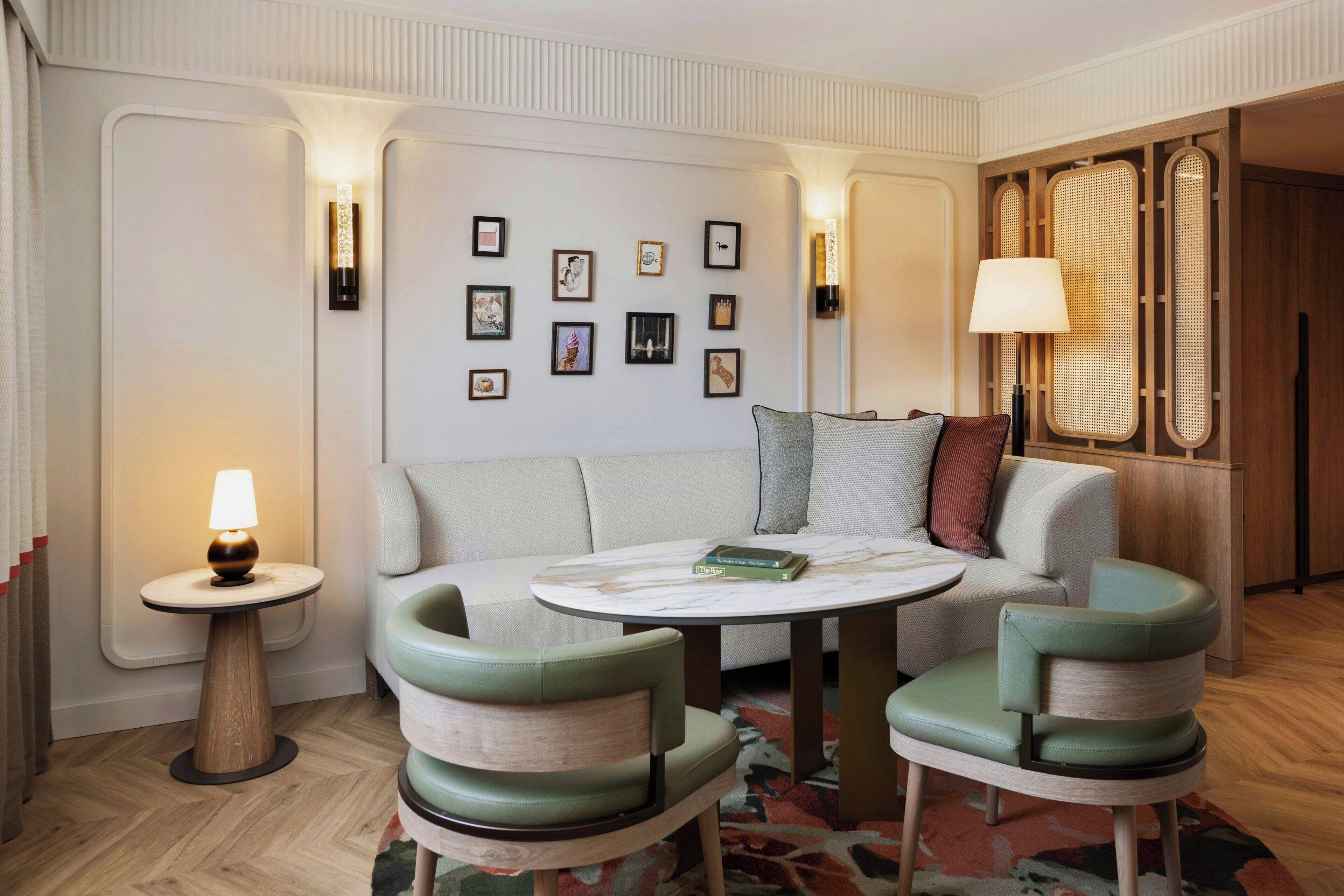 A modern hotel room with a sofa, two armchairs, a round table, and a small side table with a lamp.
