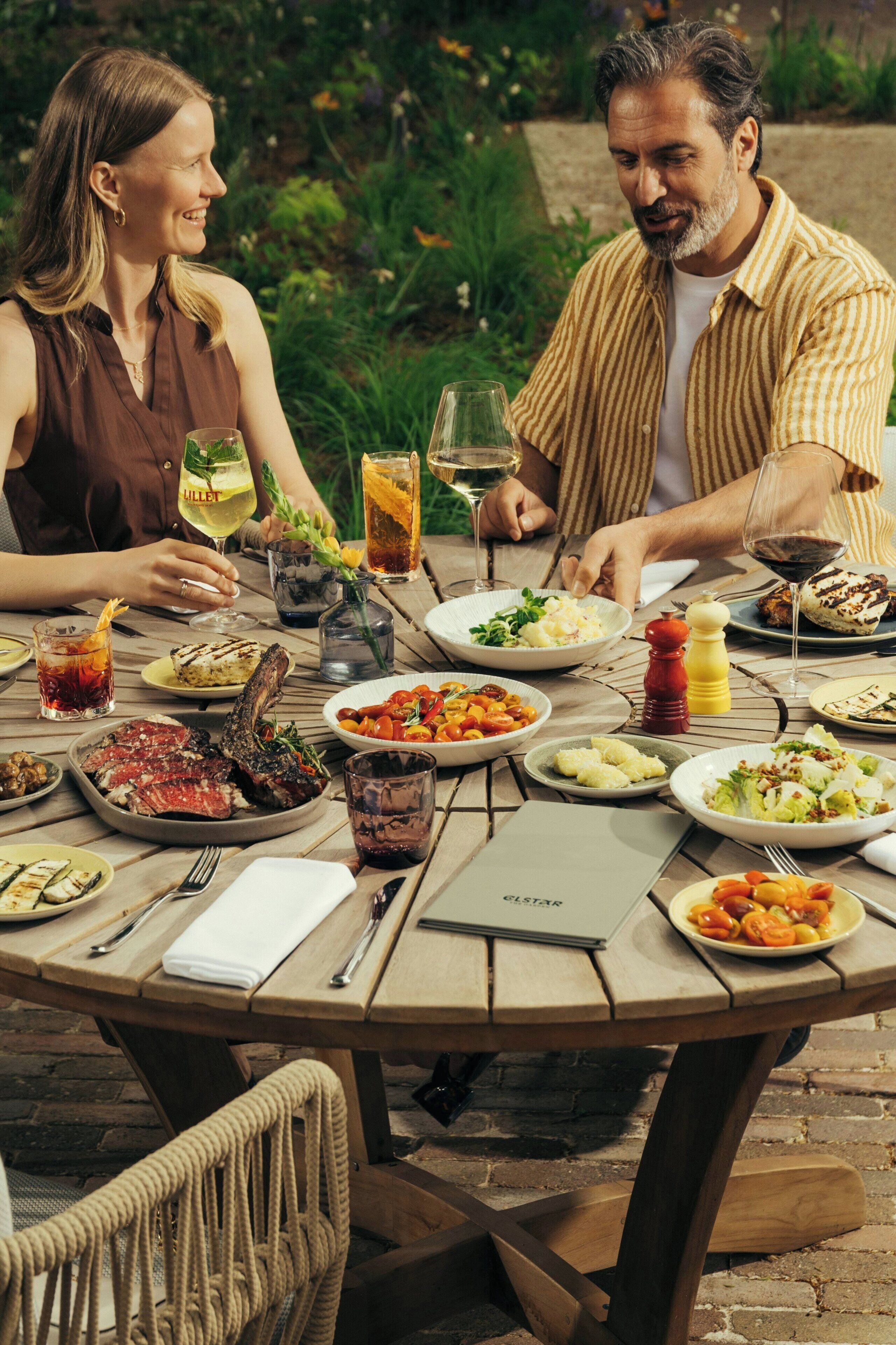 Una mesa de madera redonda con varios platos y bebidas, rodeada de sillas de mimbre.
