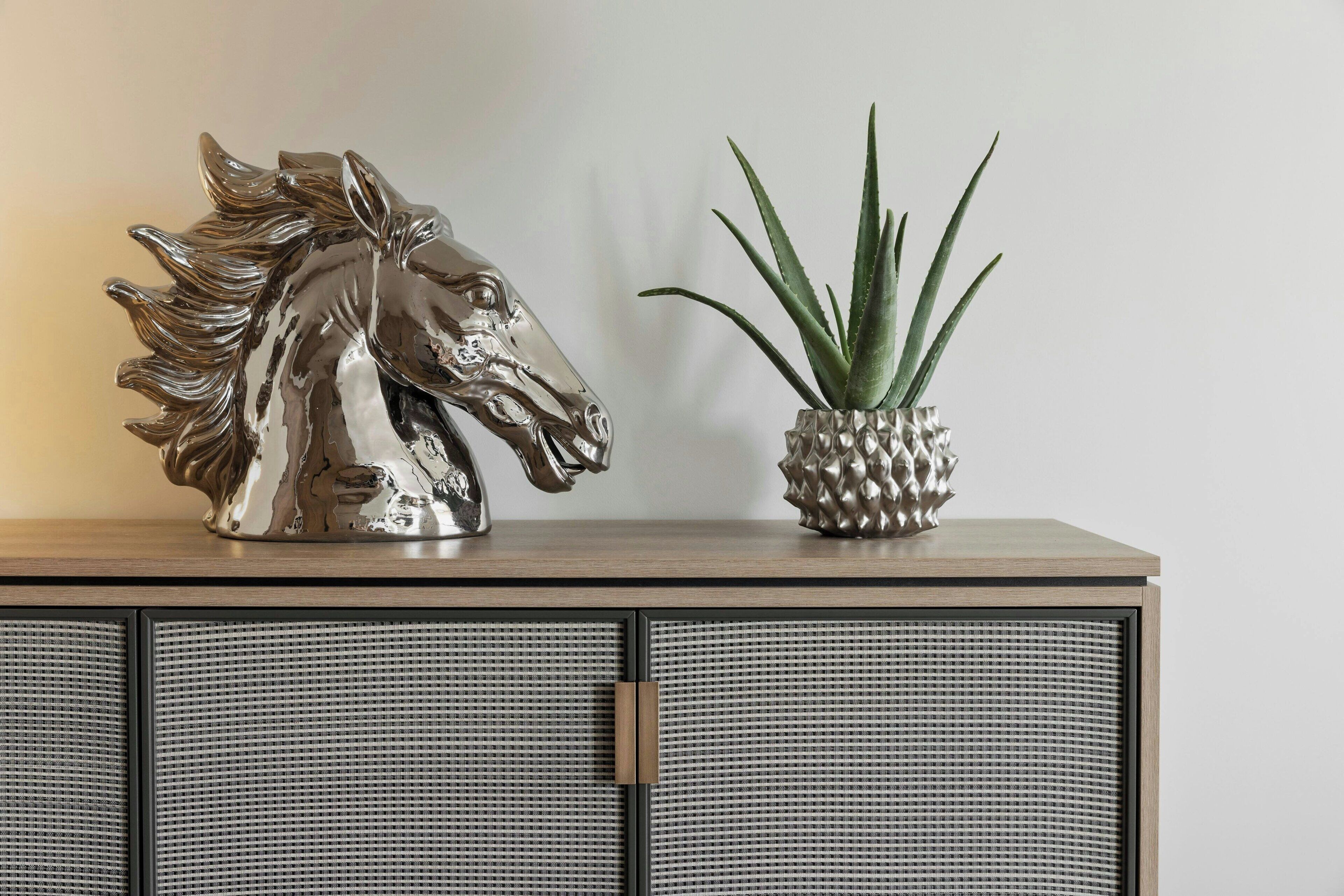 Une sculpture décorative représentant une tête de cheval et une plante en pot, posées sur un meuble en bois dont les portes sont faites d’un matériau tressé.