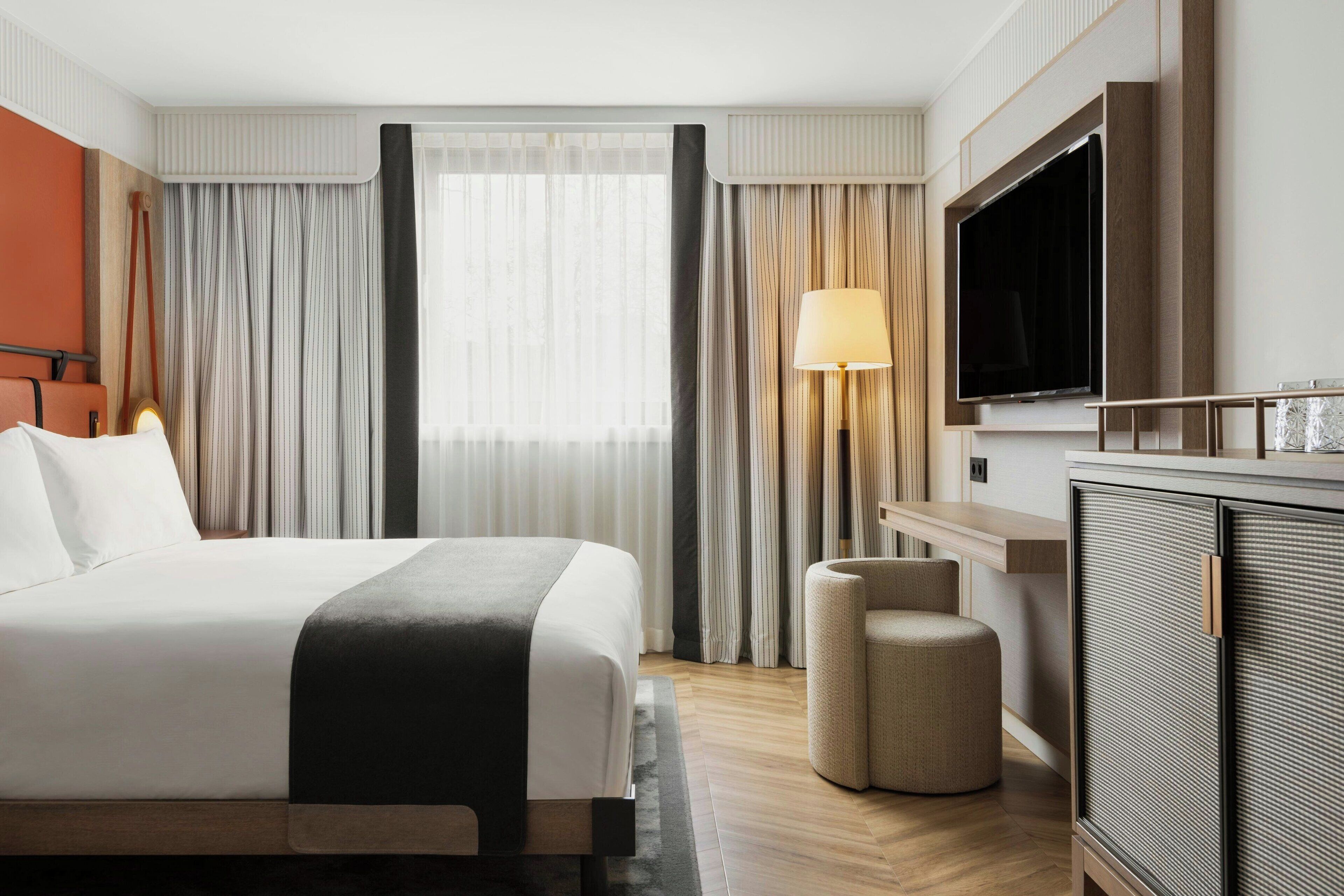 Quarto de hotel moderno com uma cama grande, televisão, escrivaninha e janela com cortinas.
