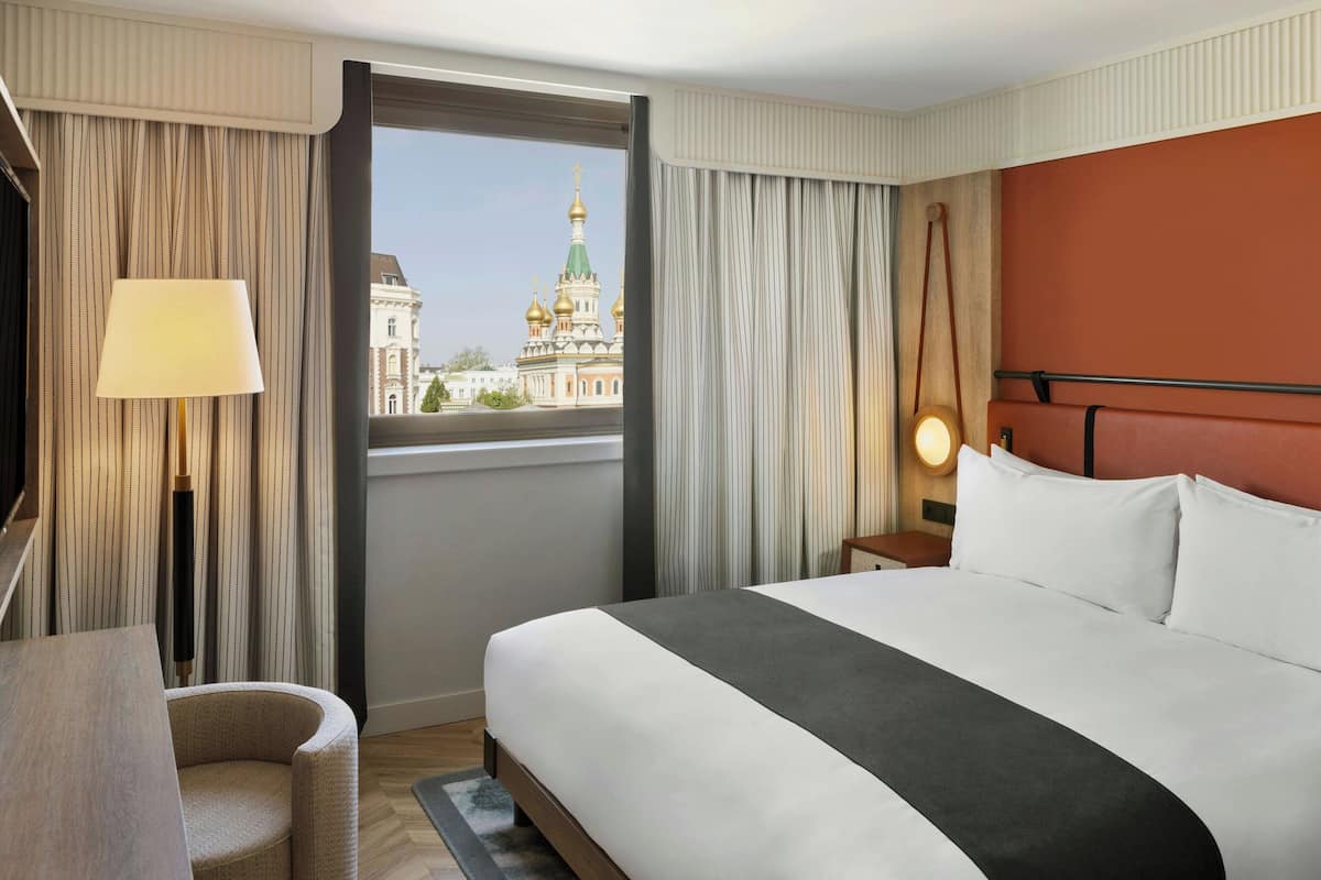 Habitación de hotel con una cama grande, un escritorio, una silla, un ventanal con vistas a un edificio y una lámpara.