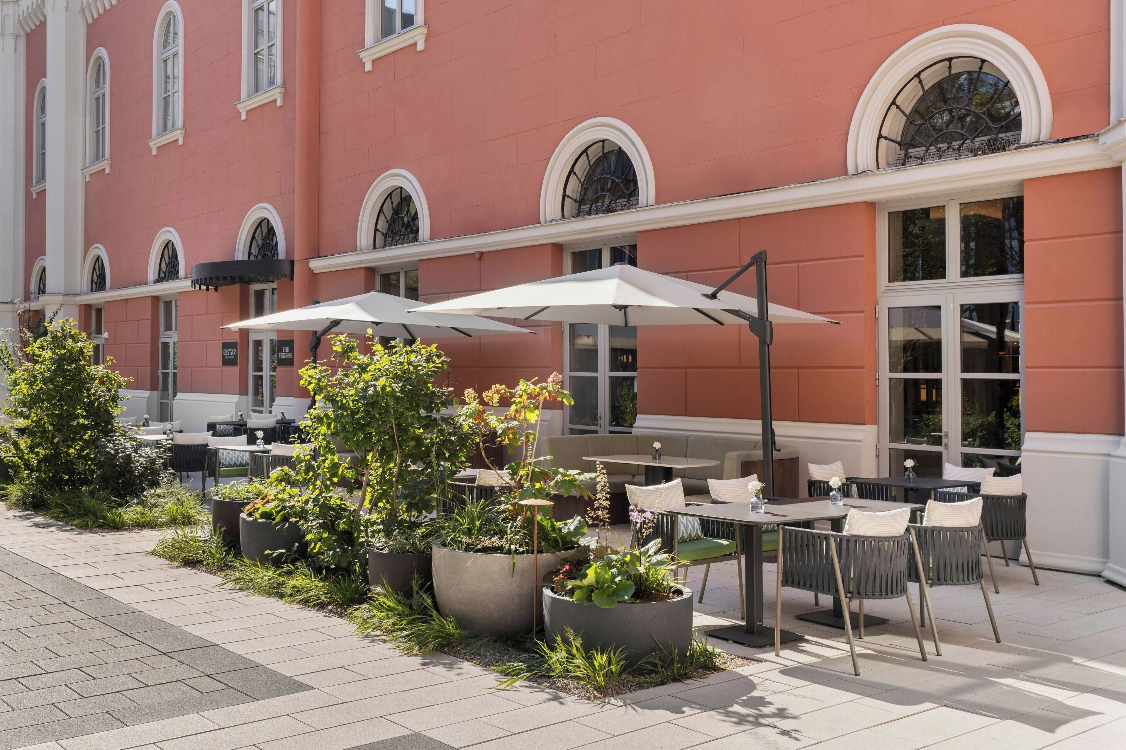 An outdoor seating area with tables and chairs, surrounded by potted plants and a building with arched windows.