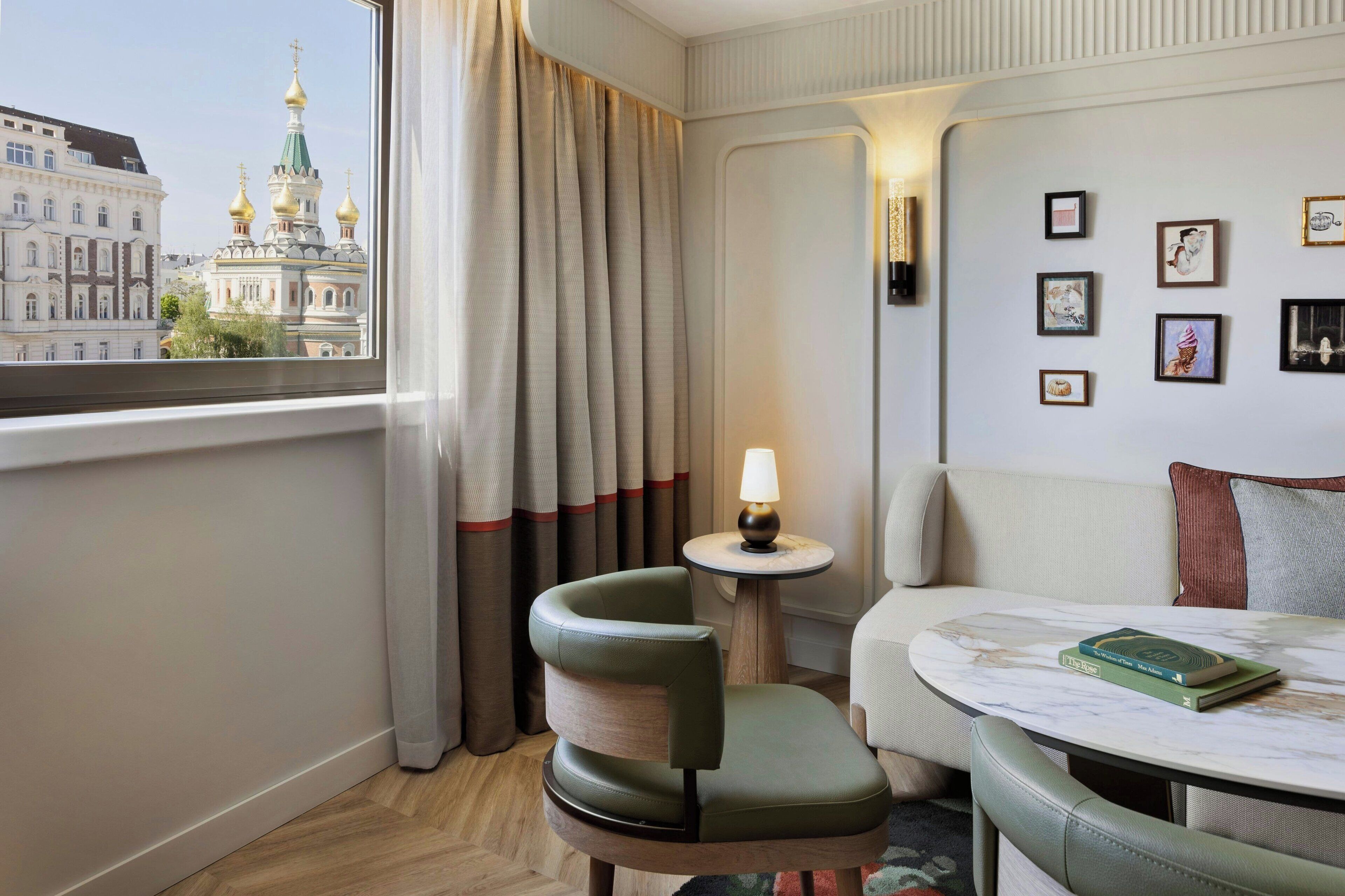 Quarto de hotel com vista para um edifício histórico, um sofá, uma cadeira, uma mesinha com uma luminária e quadros nas paredes.
