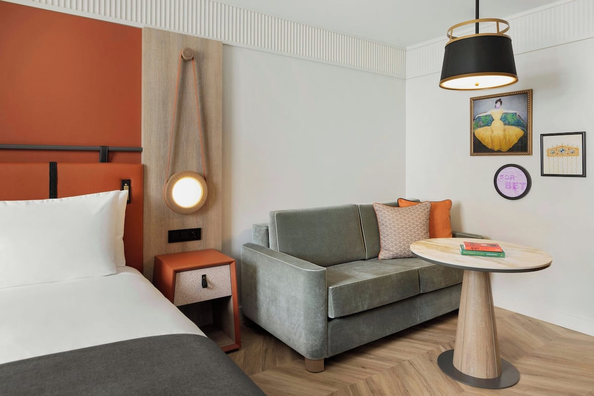 Una habitación de hotel moderna con cama, sofá gris, una mesa redonda y una obra de arte en la pared.