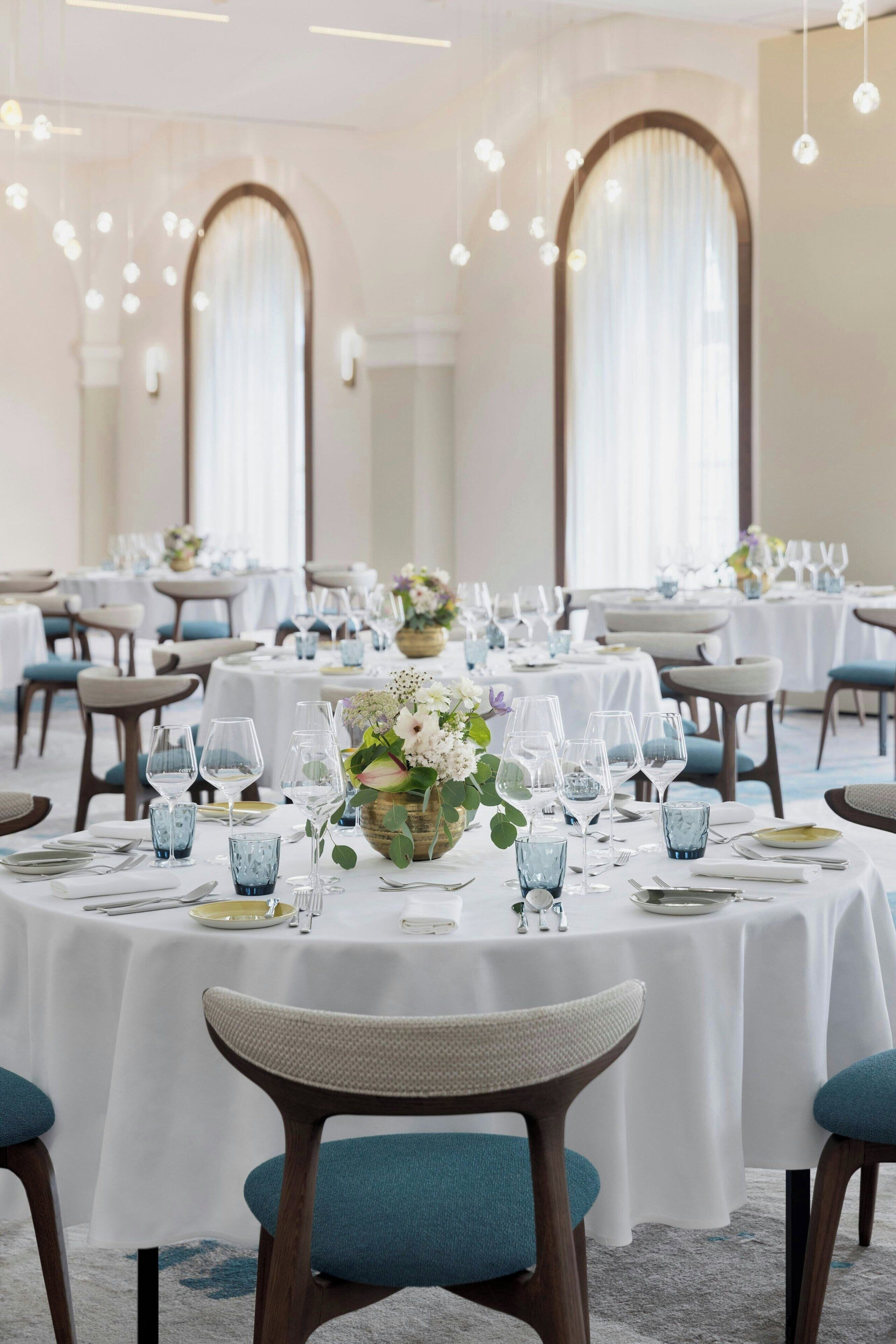Ein Festsaal mit runden Tischen, gedeckt für ein Essen, mit weißen Tischdecken, blauen Stühlen und floralen Mittelstücken.
