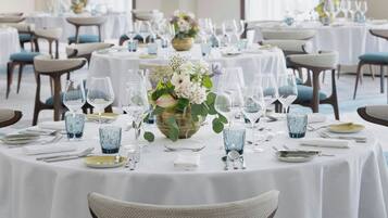 Ein Festsaal mit runden Tischen, gedeckt für ein Essen, mit weißen Tischdecken, blauen Stühlen und floralen Mittelstücken.