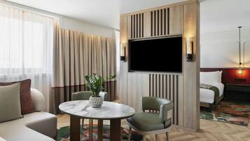Una habitación de hotel con sofá, una mesa de comedor con sillas, televisor montado en la pared, cama y mesita de noche con lámpara.