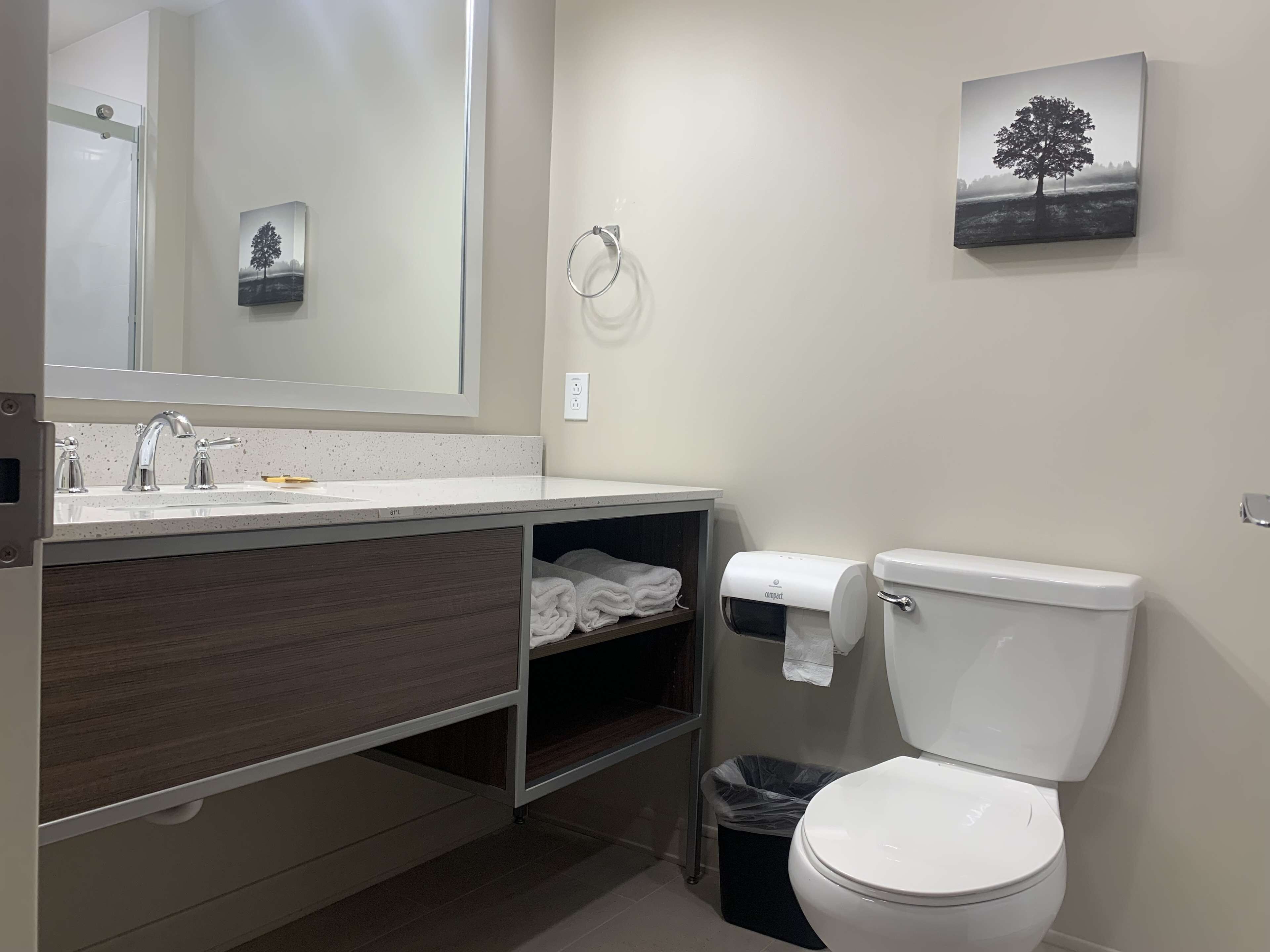 A modern bathroom with a white sink, mirror, and toilet. There are two towel racks, a trash bin, and a shelf with folded towels.