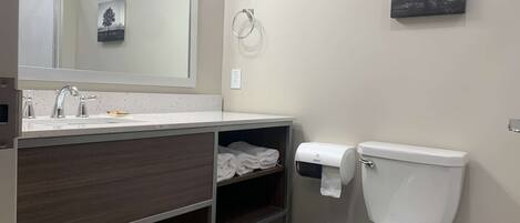Un baño moderno con lavamanos blanco, espejo e inodoro. Hay dos toalleros, un cesto de basura y un estante con toallas dobladas.