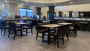 A modern restaurant with a curved ceiling, wooden tables and chairs, a coffee station, and a TV screen.