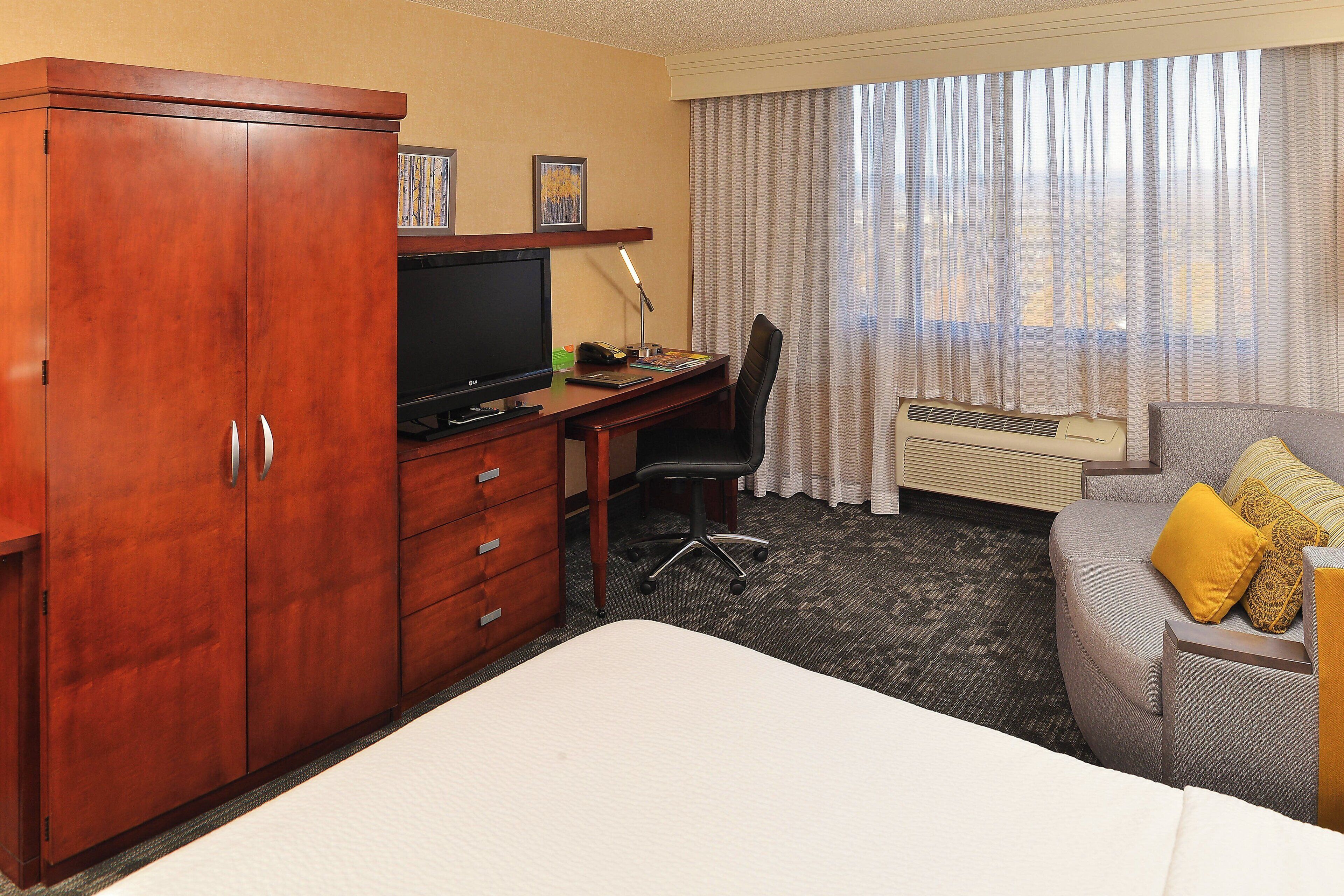 A hotel room with a wooden wardrobe, a desk with a television, a chair, a bed, and a sofa with cushions.
